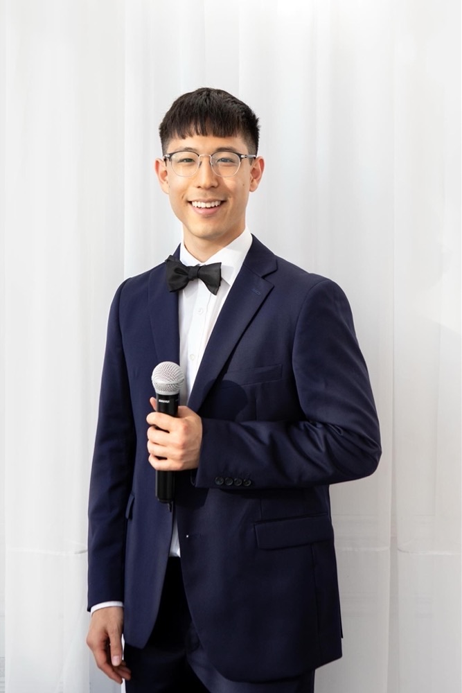 Sunwoong in a navy tuxedo holding a microphone, MC'ing a wedding in June 2024.